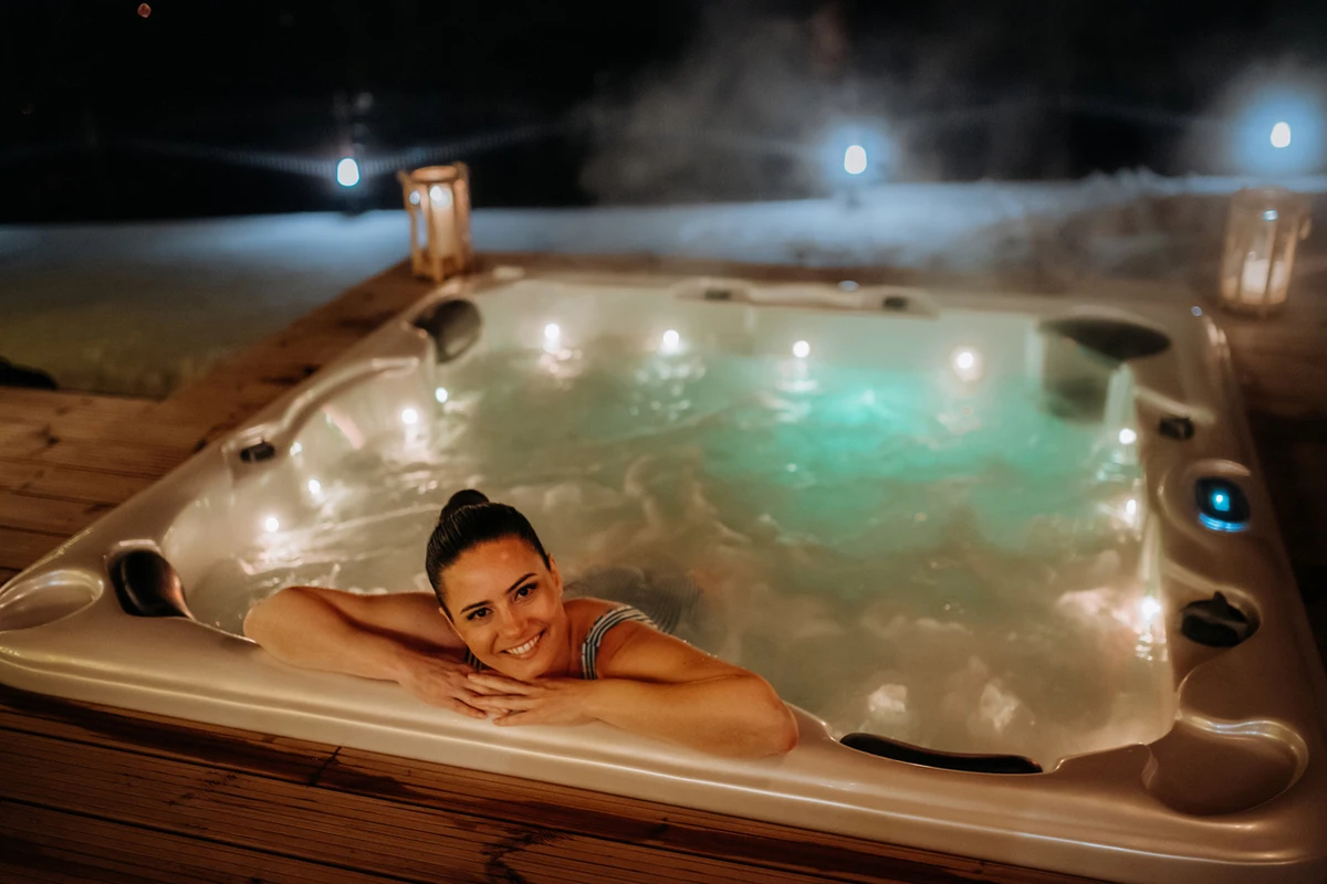 Smiling woman relaxing in a winter hot tub on an outdoor wooden deck at night, with steam rising and lanterns glowing around the spa.